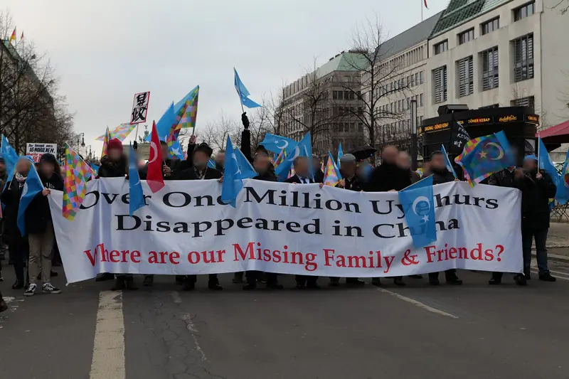 Marchers on a wide avenue carrying a long banner and pale blue East Turkestan flags.