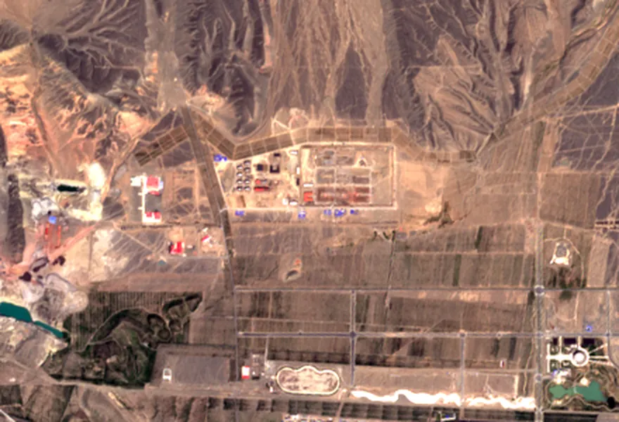 Gravel plain below bare mountains. One rectangular boundary line encloses earthworks and a few small buildings; around it there is bare ground, tracks and ploughed strips.
